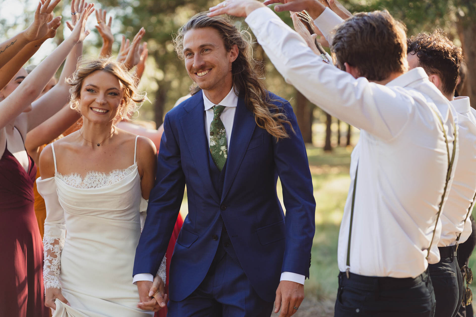 A bride and groom walk hand in hand through a celebratory arch of raised hands surrounded by wedding guests among Ponderosa pines in Flagstaff, Arizona